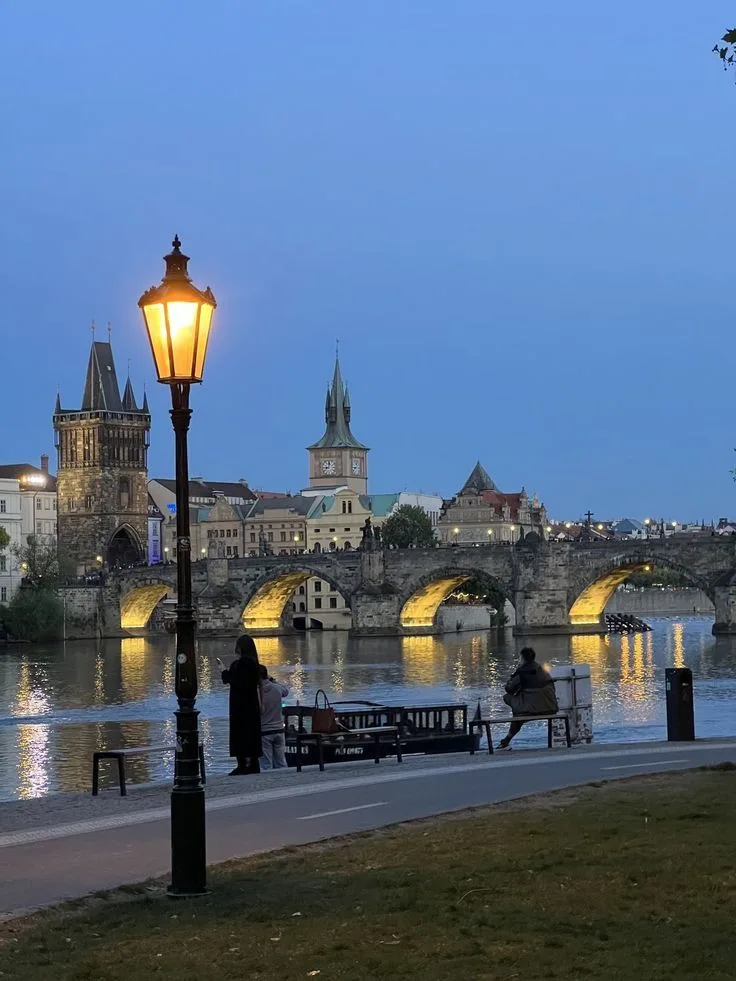 Charles Bridge and the Old Town skyline at blue hour, lit by a riverside lamp